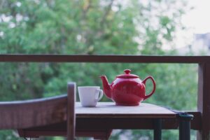 selective focus photo of red ceramic teapot and white ceramic teacup on brown wooden table near tree during daytime